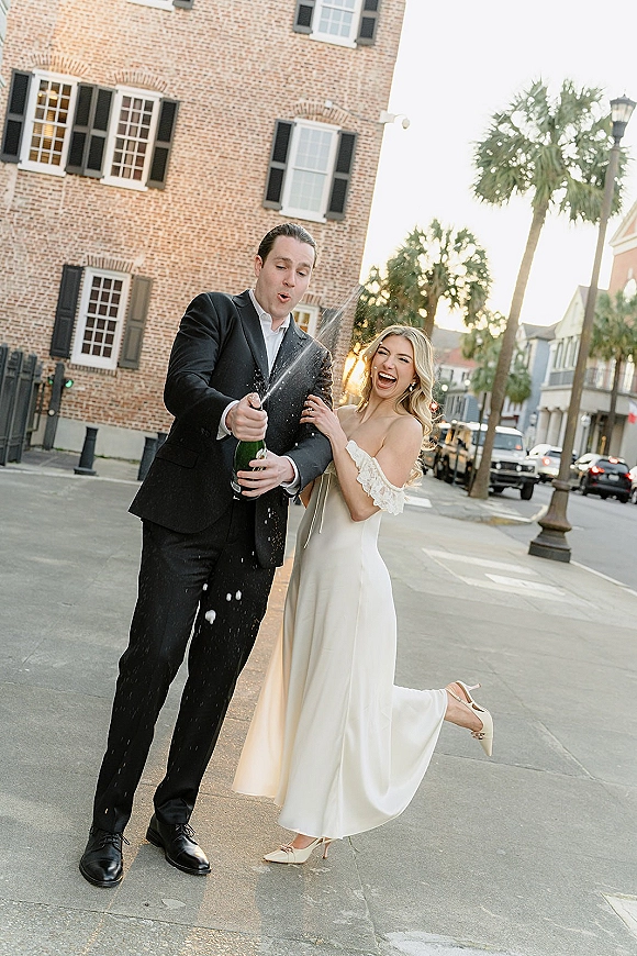 Champagne pop moment as bride and groom laugh while champagne spray bursts on a city sidewalk at sunset, brick storefronts behind