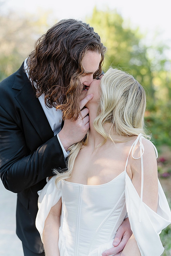 Wedding kiss portrait of bride in off-shoulder corset gown and groom in black tuxedo embracing on a garden path with greenery