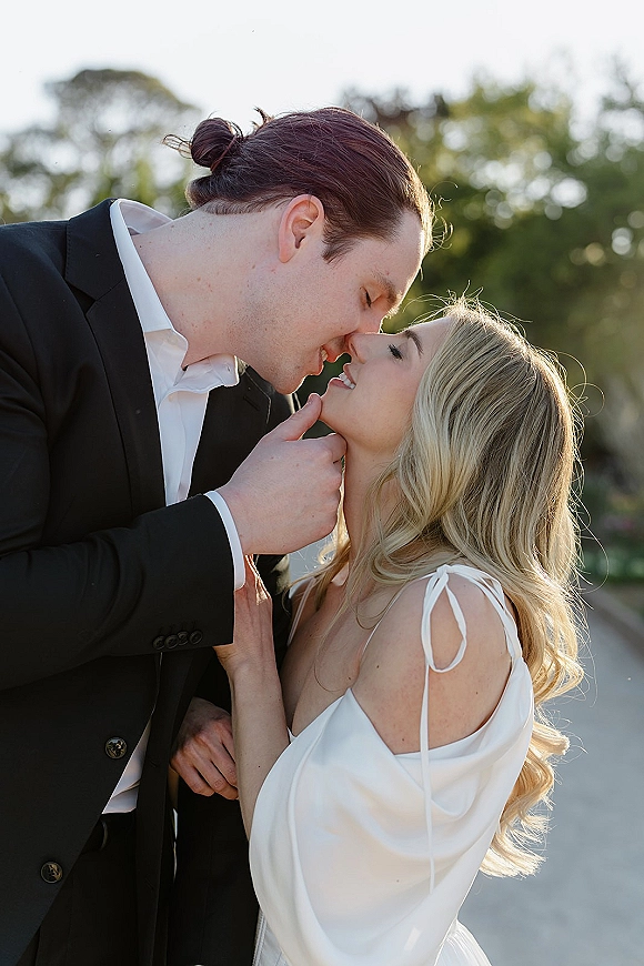 Wedding kiss portrait of bride and groom kissing as he holds her chin, off-shoulder dress and black suit against outdoor trees at golden hour