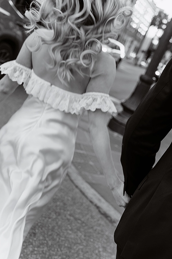 Wedding couple walking hand in hand, bride in an off-the-shoulder gown with lace sleeves, strolling down a city sidewalk with streetlight bokeh