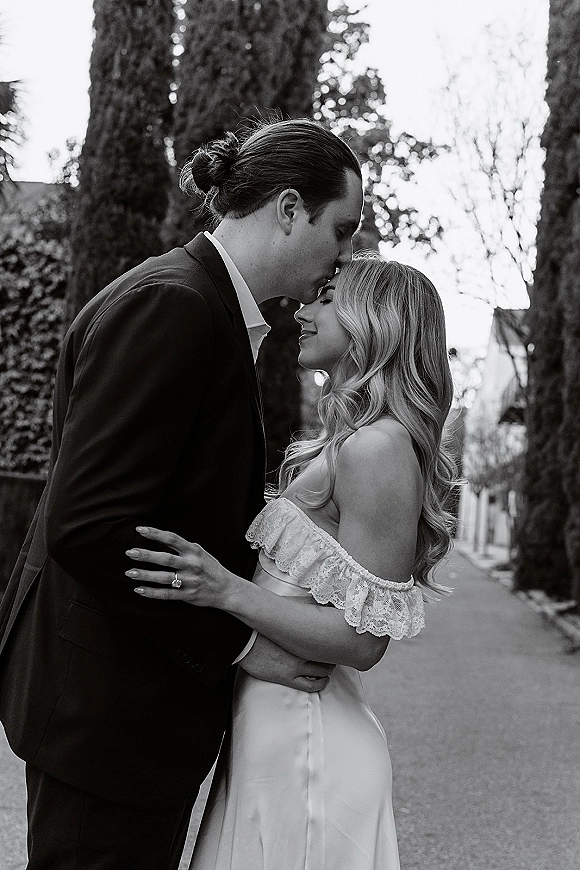 Wedding couple portrait in black and white as the groom in a black suit kisses the bride’s forehead on a tree-lined street walkway