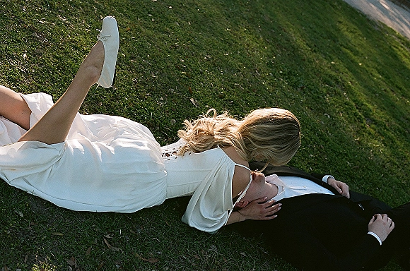 Wedding kiss as the couple lying on grass, bride in off-shoulder dress and white bridal flats leaning over groom in a black suit