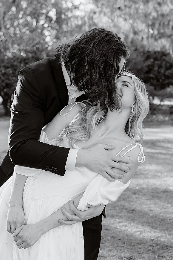 Wedding kiss portrait of groom in tuxedo and bride in spaghetti strap dress in a romantic dip, with soft garden bokeh behind them
