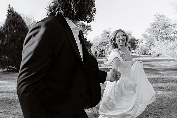 Couple portrait of bride and groom holding hands, bride twirling her wedding dress as they walk on a lawn with trees and open sky