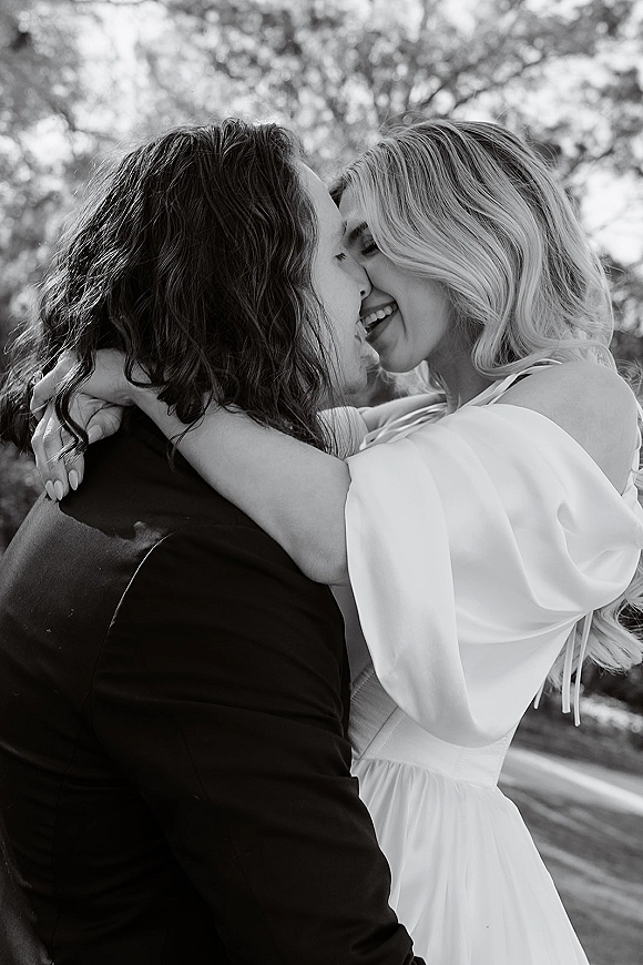 Wedding kiss portrait in a black and white wedding photo, close-up of bride and groom embracing in trees, her off-shoulder dress visible