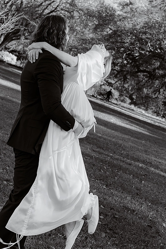Couple portrait in an outdoor wedding portrait, groom holding bride in a dip as her long sleeve gown flows on a tree-lined lawn