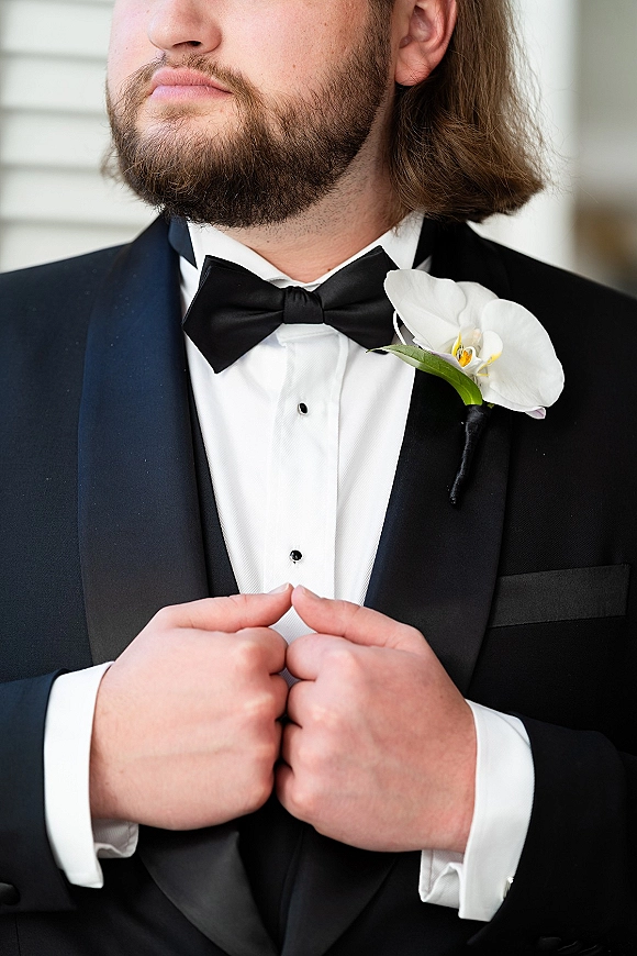 Groom portrait in a black tuxedo with satin lapels and black bow tie, wearing a white orchid boutonniere against a light wall