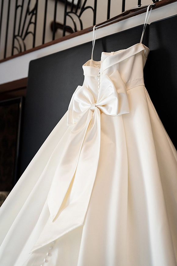 Wedding dress, satin wedding dress hanging on a hanger with a button back and satin bow near a staircase railing indoors