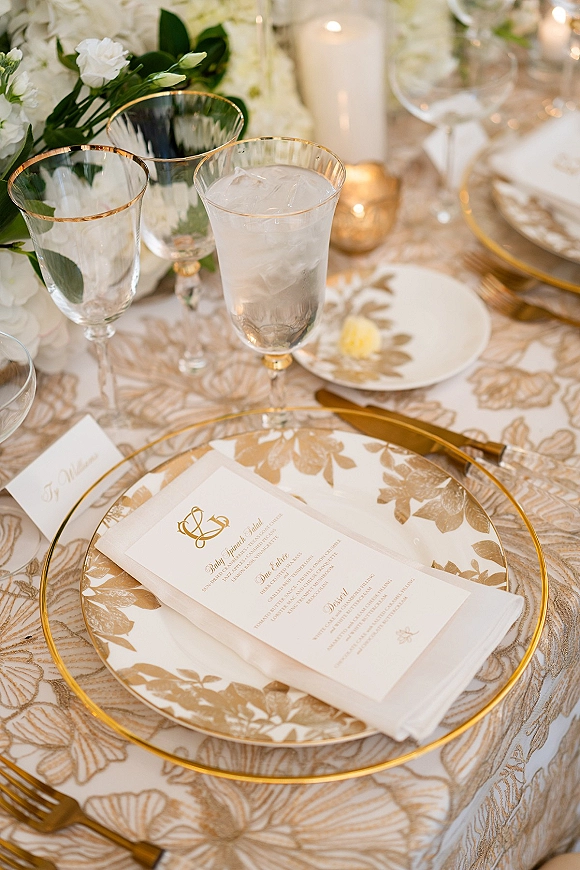 Reception tablescape with gold and white tablescape details, gold rim plates, patterned dinner plates, menu card, candles and white florals amid bokeh lights