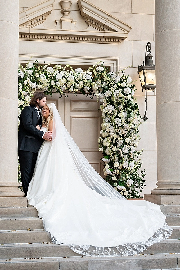 Wedding couple portrait of bride and groom embrace with a forehead kiss on stone steps under a white floral arch at a grand doorway