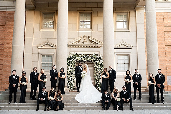 Wedding party portrait of bride and groom with wedding party on stone steps, framed by a white floral arch at a columned building facade