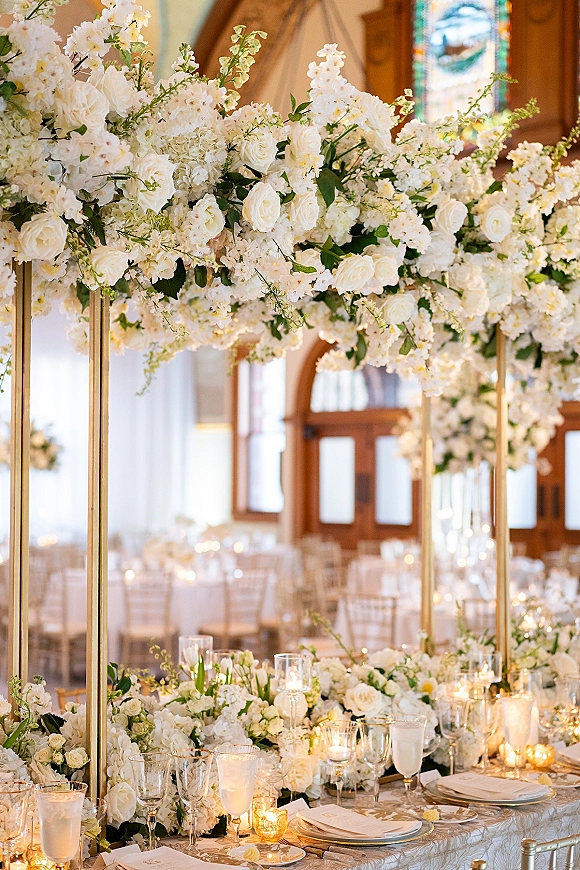 Reception tablescape with wedding head table decor, white roses and hydrangea under floral canopy on gold stand, candlelit by stained glass window