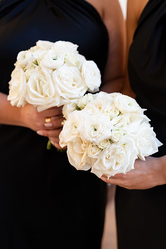 Bridesmaid bouquets of white roses and ranunculus held against black dresses, with a ring accent in a neutral indoor setting