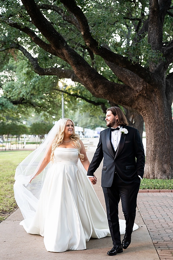 Couple portrait of bride and groom walking hand in hand under a tree canopy, her cathedral veil trailing beside his black tuxedo