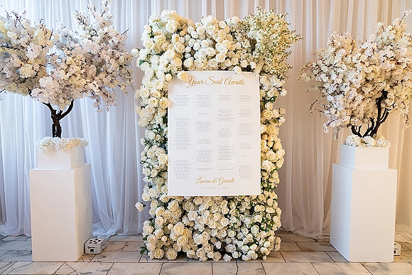 Seating chart display with wedding seating chart calligraphy on a sign, framed by white rose floral wall and cherry blossom trees before drapes