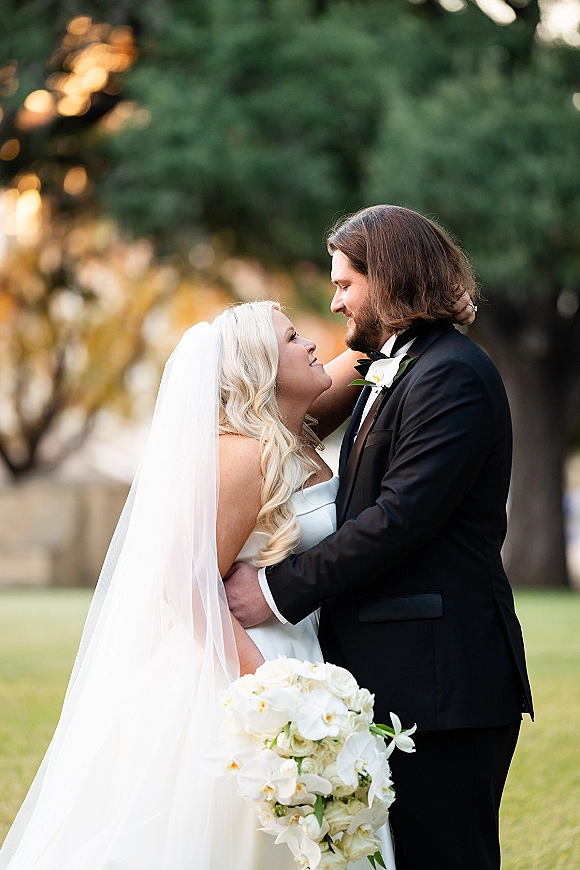 Couple portrait of bride and groom embracing, bride looking up at him in veil and strapless dress, holding white orchid bouquet on lawn by trees