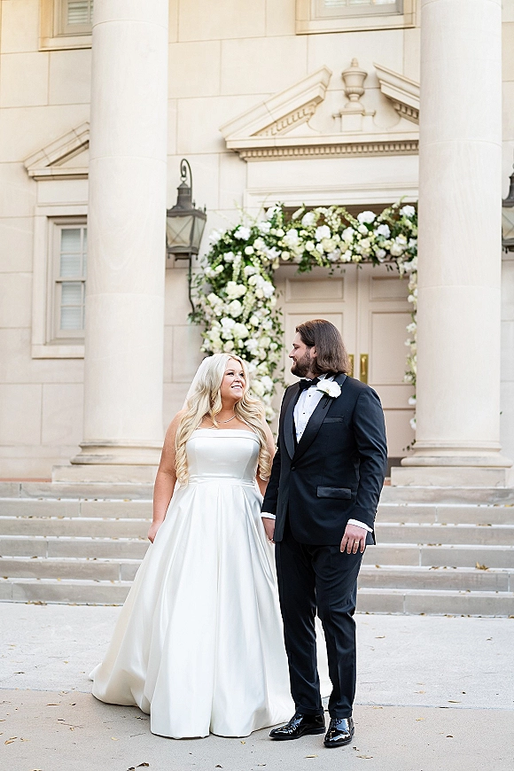 Couple portrait of bride and groom holding hands, gazing at each other on stone steps by white columns beneath a floral arch