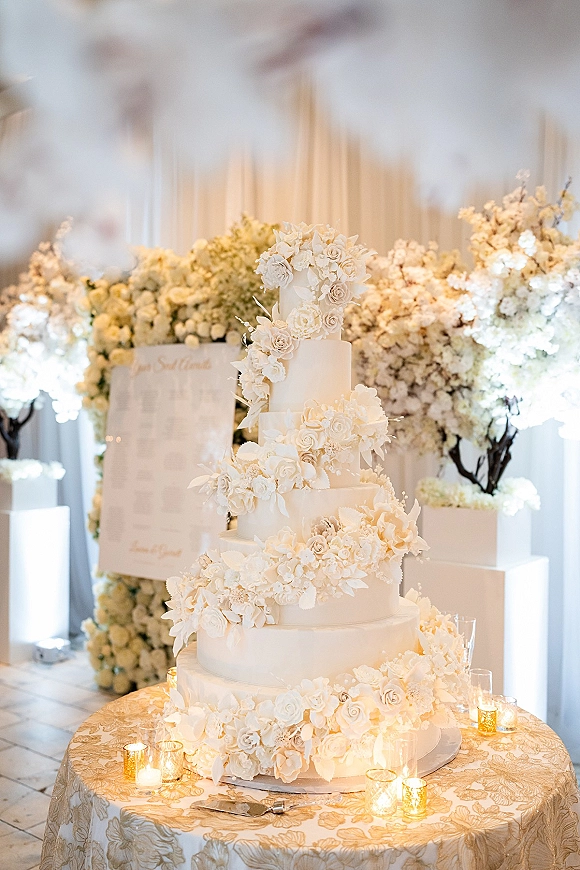 Wedding cake with tiered white design and cascading sugar flowers on a stand, surrounded by votive candles and a floral wall backdrop
