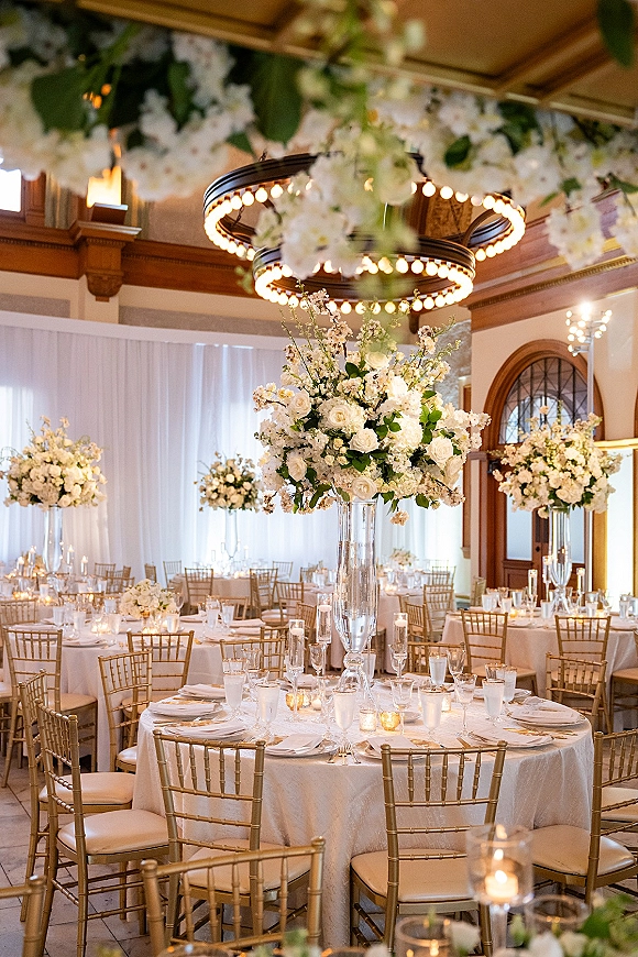 Reception tablescape with round wedding tables, tall white rose and hydrangea centerpieces in glass trumpet vases beneath chandeliers