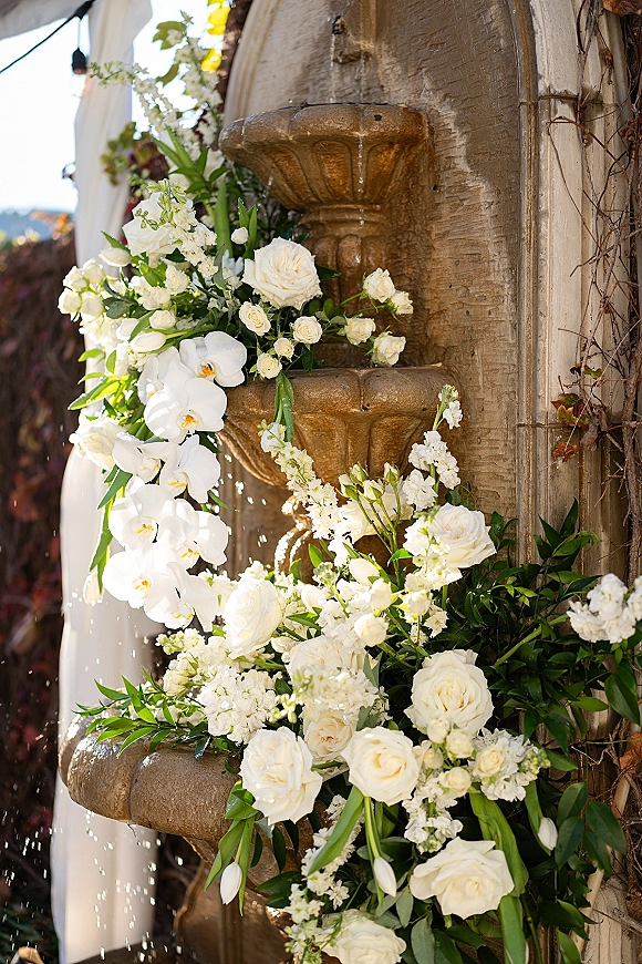 Wedding fountain decor with white roses and orchids, tulips, and greenery garland draped over a stone fountain against an ivy wall