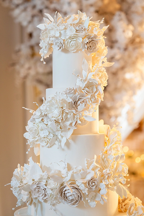 Wedding cake with white fondant tiers and cascading sugar flowers—roses and peonies—set against a blurred floral backdrop and warm reception lights