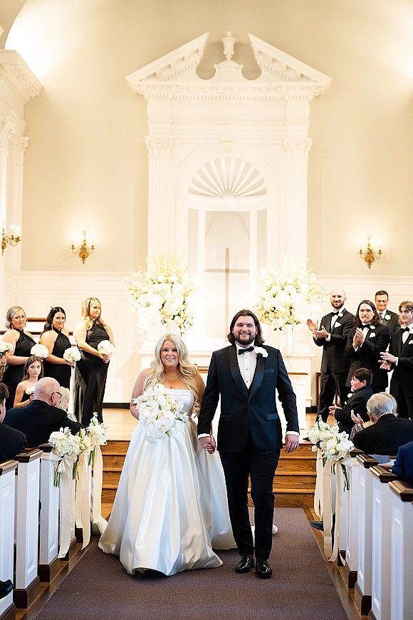 Wedding recessional as bride and groom walk the church aisle holding hands, guests clapping, with bouquets and aisle flowers with ribbon accents