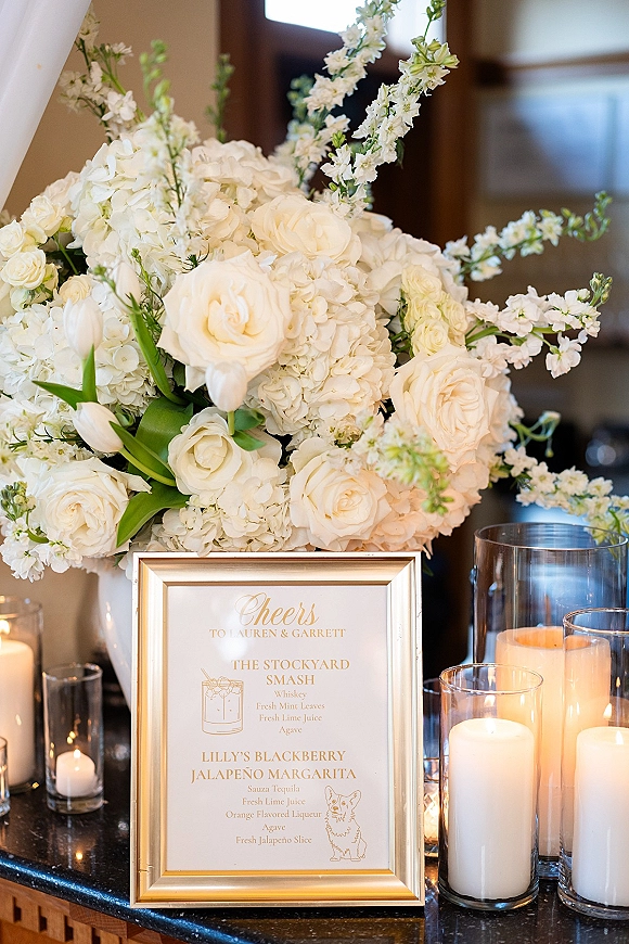 Wedding bar sign and signature cocktail sign in a gold frame beside white roses and candlelit hurricane vases on an indoor bar counter