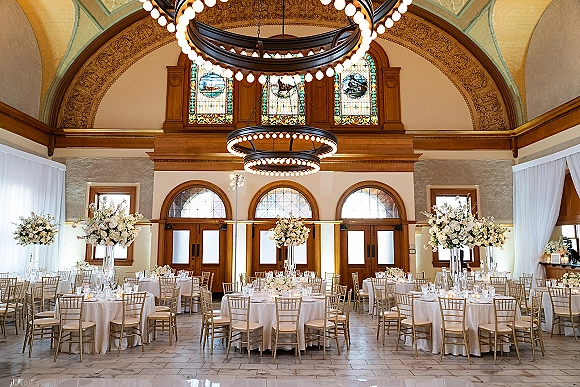 Reception tablescape with white linens on round tables, gold Chiavari chairs, tall white floral centerpieces, and votive candles in a chandelier ballroom