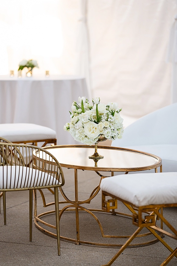 Wedding lounge seating with white floral centerpiece in a gold pedestal bowl on a glass coffee table inside a white drape tent