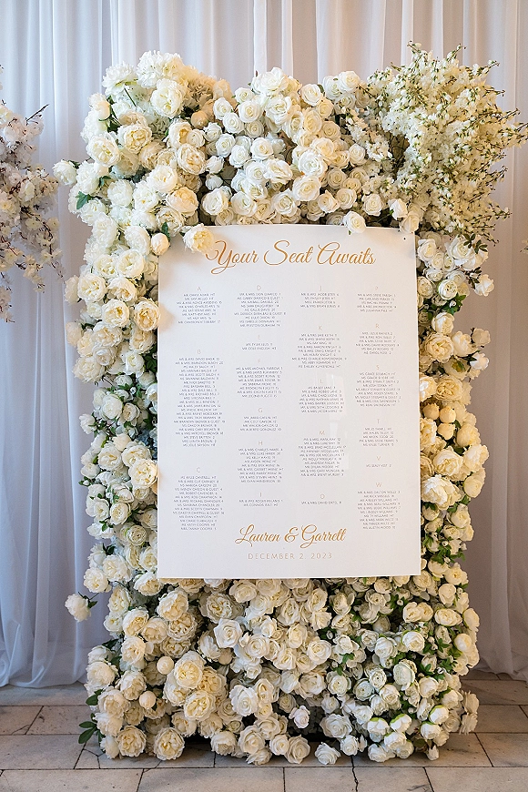 Wedding seating chart sign with calligraphy lettering framed by white roses, peonies, and greenery against a white curtain backdrop