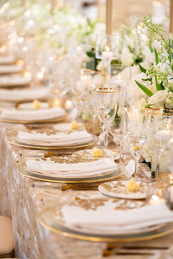 Reception tablescape with wedding place setting, gold-rim charger plates, crystal wine glasses, and candlelit white florals on lace tablecloth﻿