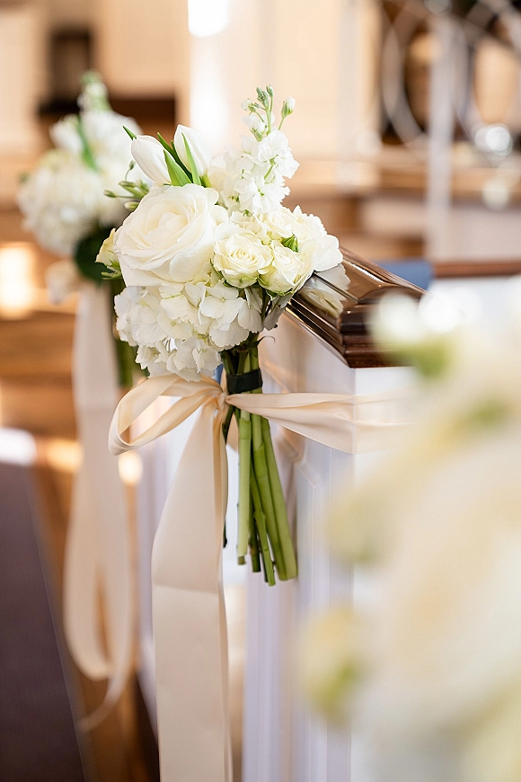 Ceremony aisle flowers with white roses and hydrangea on a pew clip, tied with an ivory ribbon bow along a church aisle runner