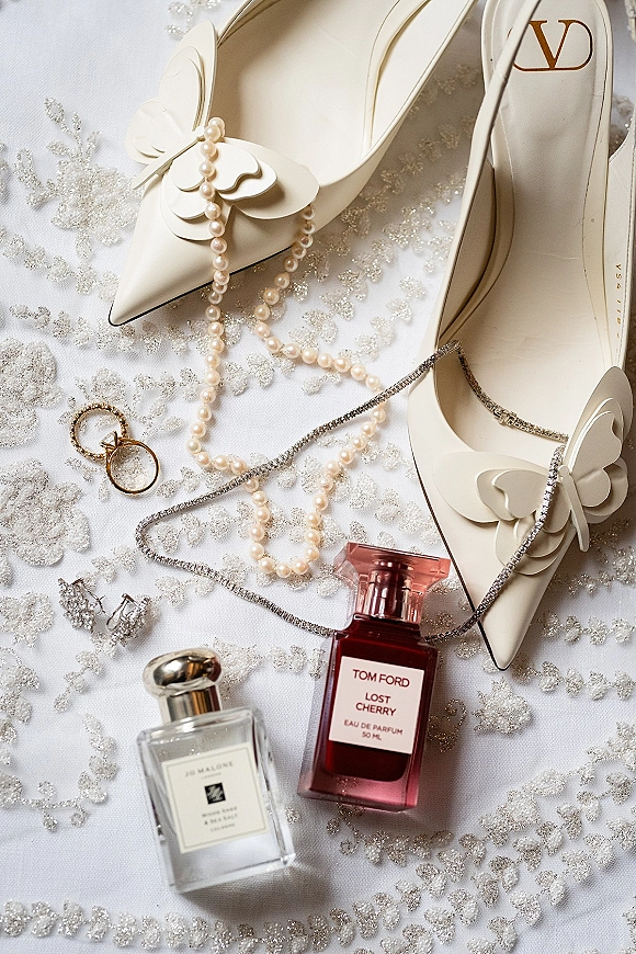 Bridal shoe flatlay with ivory wedding heels featuring butterfly appliques, pearl jewelry, rings, and perfume on white fabric backdrop