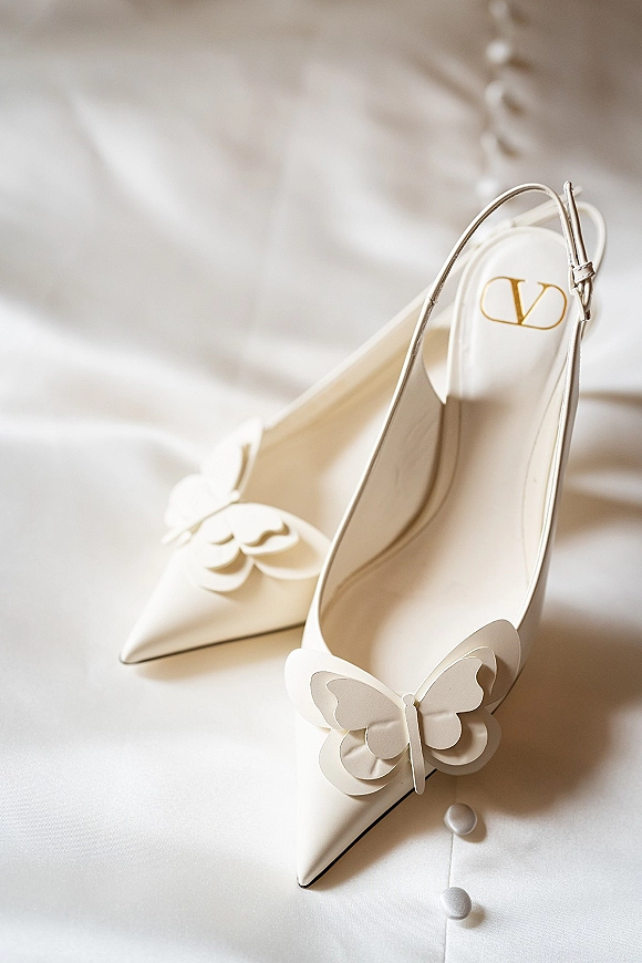 Bridal shoes, white wedding heels with butterfly appliques, arranged on white fabric beside a row of dress buttons in a flat lay