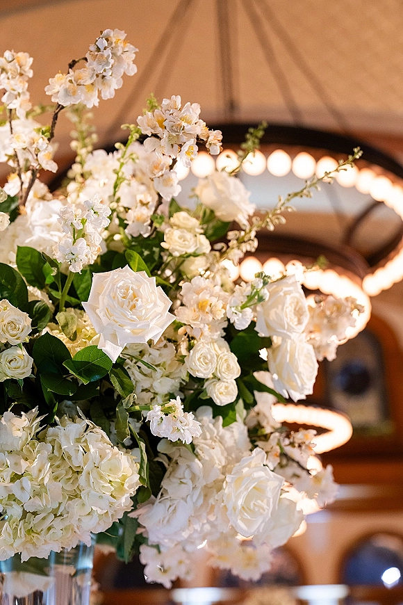 Wedding floral centerpiece with white roses and hydrangea in a tall glass vase, greenery and branches under warm string lights indoors