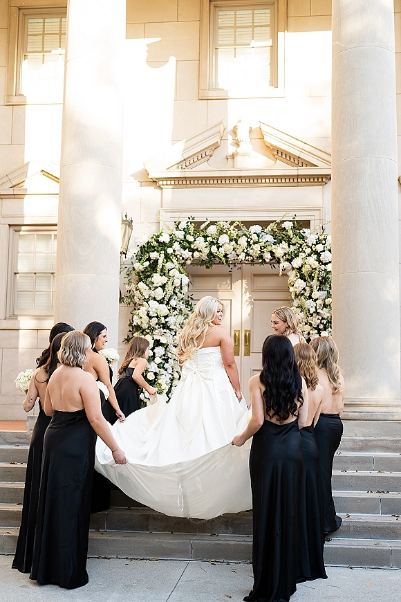 Bride with bridesmaids as bridesmaids helping bride hold her long dress train, each carrying white bouquets on classic building steps by a floral arch