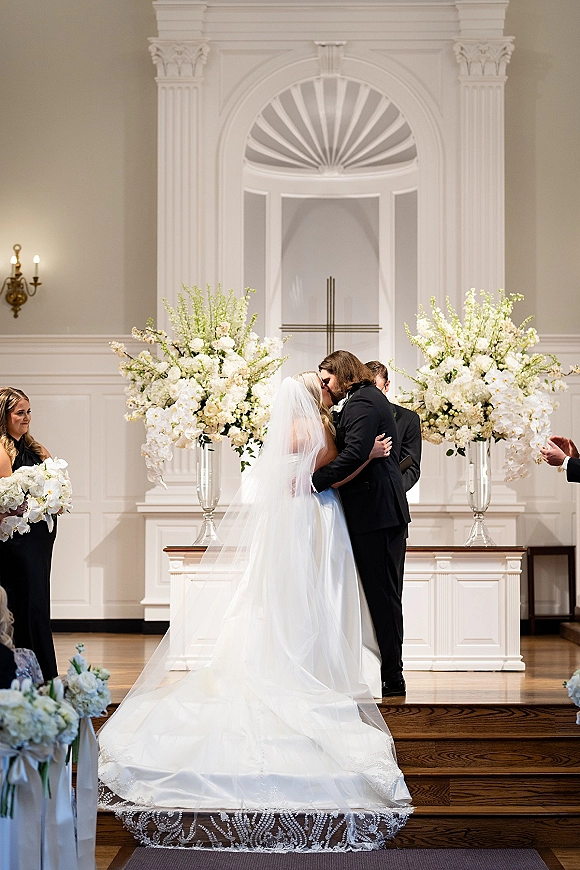 Wedding kiss as bride and groom embrace at a church altar, her long veil and dress train flowing beneath a cross and white flowers