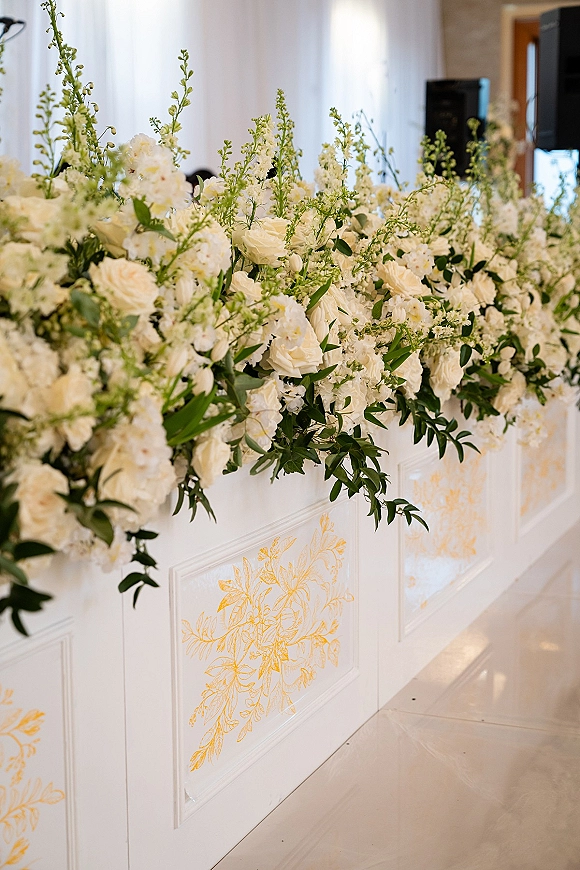 Wedding floral installation with white floral garland and white roses over a white draped, gold-detailed bar front in an indoor reception space