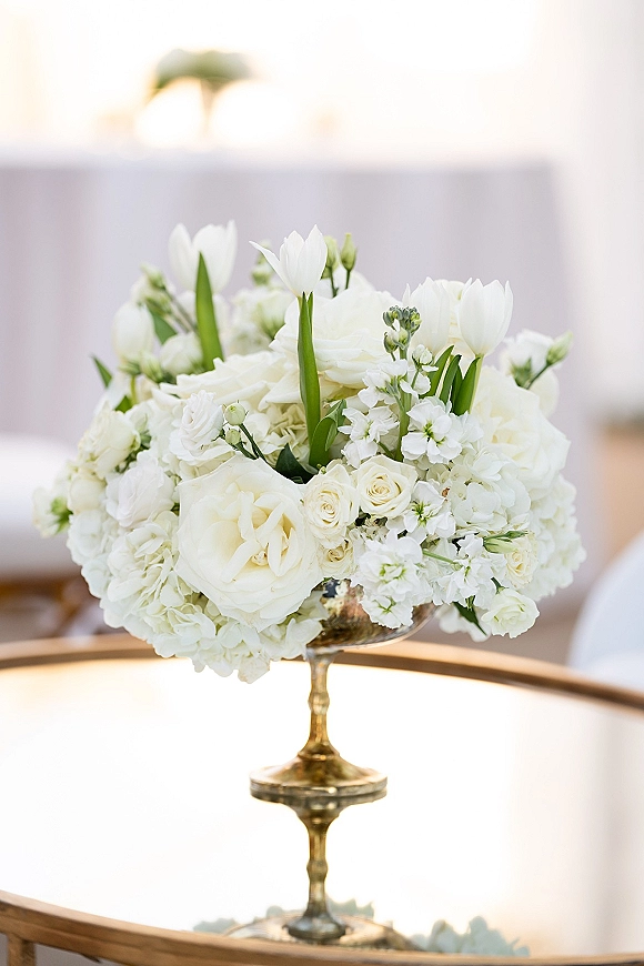 Wedding centerpiece with a white floral centerpiece of roses, tulips, and hydrangea in a gold compote vase on a round mirror base