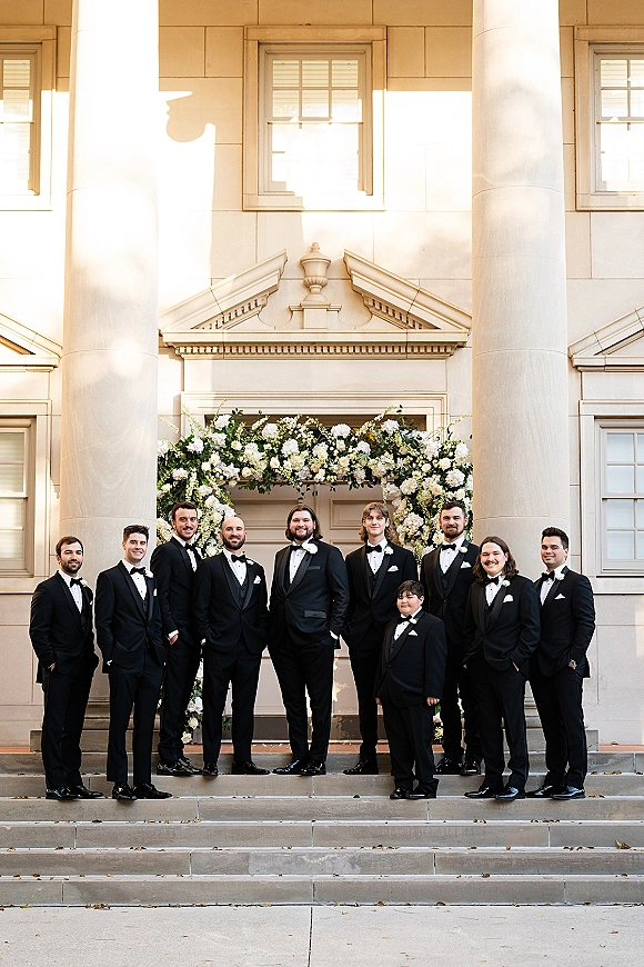 Groomsmen portrait of men in black tuxedos with bow ties and boutonnieres standing by a white floral arch on columned building steps