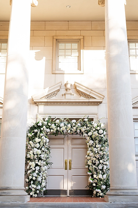 Floral ceremony arch of white flowers and greenery framing double doors on a columned white facade, creating an elegant wedding entrance backdrop