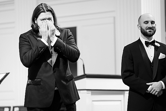 Groom emotional moment as he wipes tears in a black tuxedo with boutonniere, standing by a podium in an indoor ceremony space
