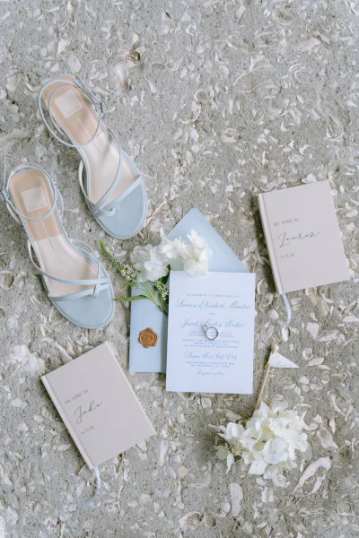 Wedding invitation suite with dusty blue wedding invitations, paired with linen vow books on a stone surface dotted with seashell fragments.