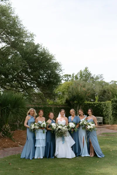 Bride and bridesmaids in dusty blue bridesmaid dresses pose together on a garden lawn framed by trees and hedges under an open sky.