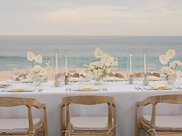 Reception tablescape with white linens, calla lily centerpieces, and glass hurricane candles on a long table overlooking the ocean horizon