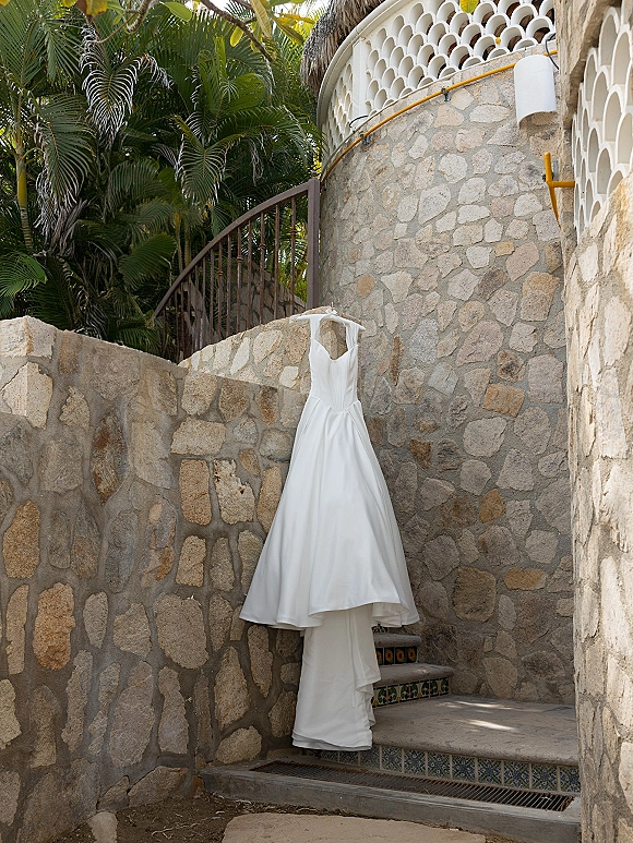 Wedding dress in satin, spaghetti straps on a hanger against a stone wall by outdoor stairs with wrought iron gate and palms