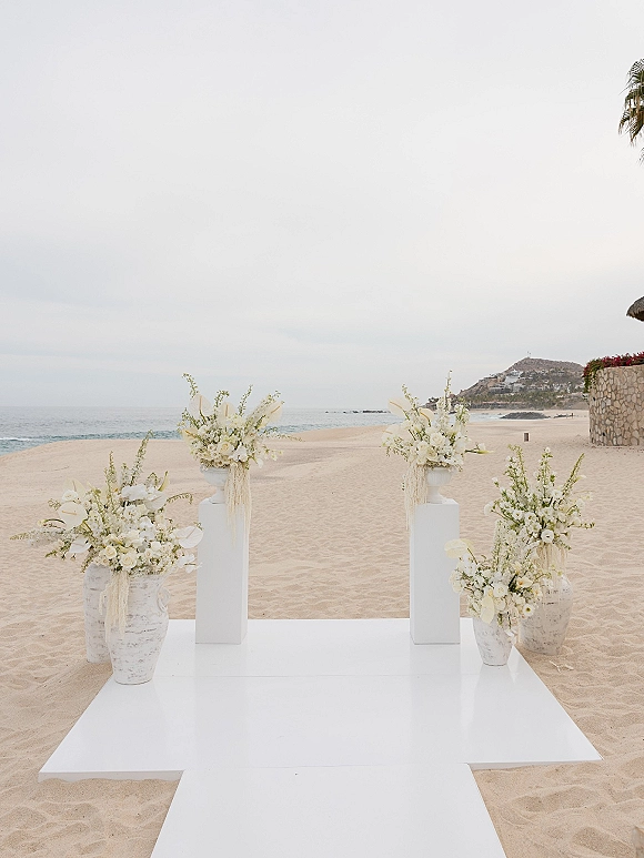 Beach ceremony setup with an oceanfront wedding ceremony white platform aisle, ceramic vases and white florals on a sandy shore