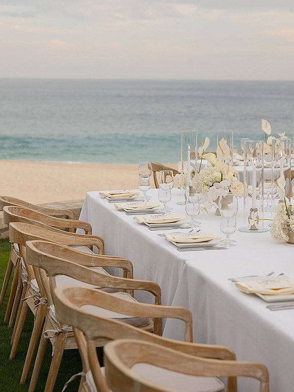 Reception tablescape with white linens, wooden chairs, menus and glassware, plus tall cylinder candles and white floral centerpiece by the ocean backdrop