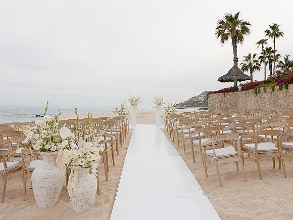Beach ceremony setup with an oceanfront wedding aisle, white aisle runner on sand lined by wooden chairs and urn florals by the sea