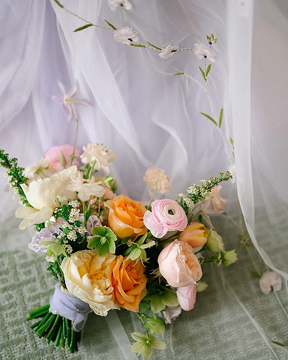 Bridal bouquet flatlay with pastel wedding bouquet blooms—garden roses, ranunculus, and greenery—wrapped with ribbon on linen backdrop